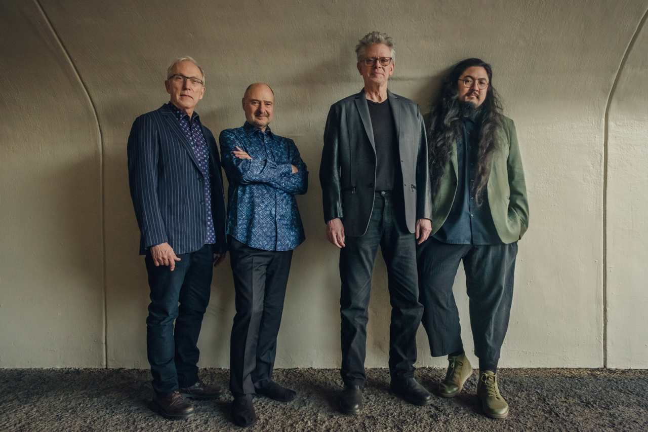 Kronos Quartet - four men posing against a beige wall looking directly at the camera