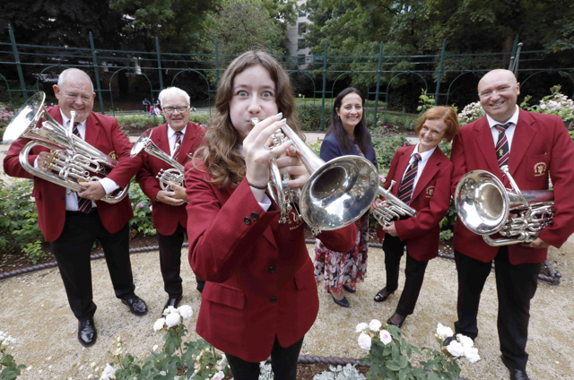 Men and women dressed in wine blazers with brass instruments in the rose garden in the Iveagh Gardens posing with Minister Catherine Martin TD in a Music Network photocall